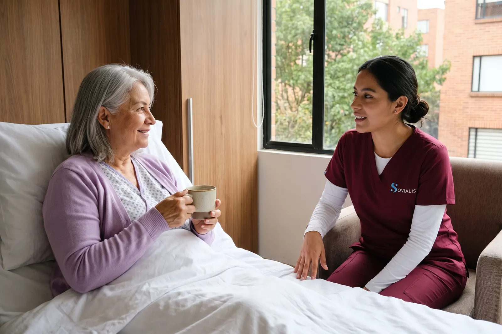 Cuidadora hospitalaria sentada junto a la cama de una persona mayor en una habitación de clínica en Bogotá