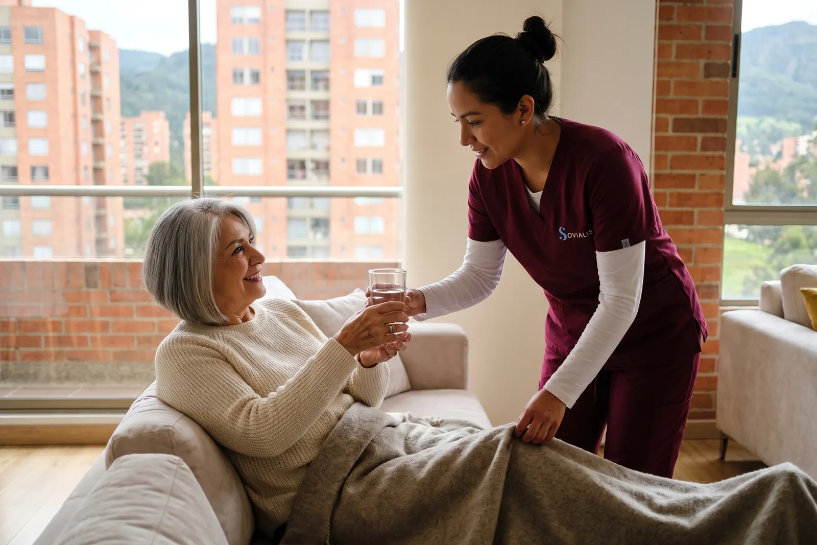 Cuidadora acerca una manta y un vaso de agua a una señora mayor que se recupera en el sofá de su apartamento