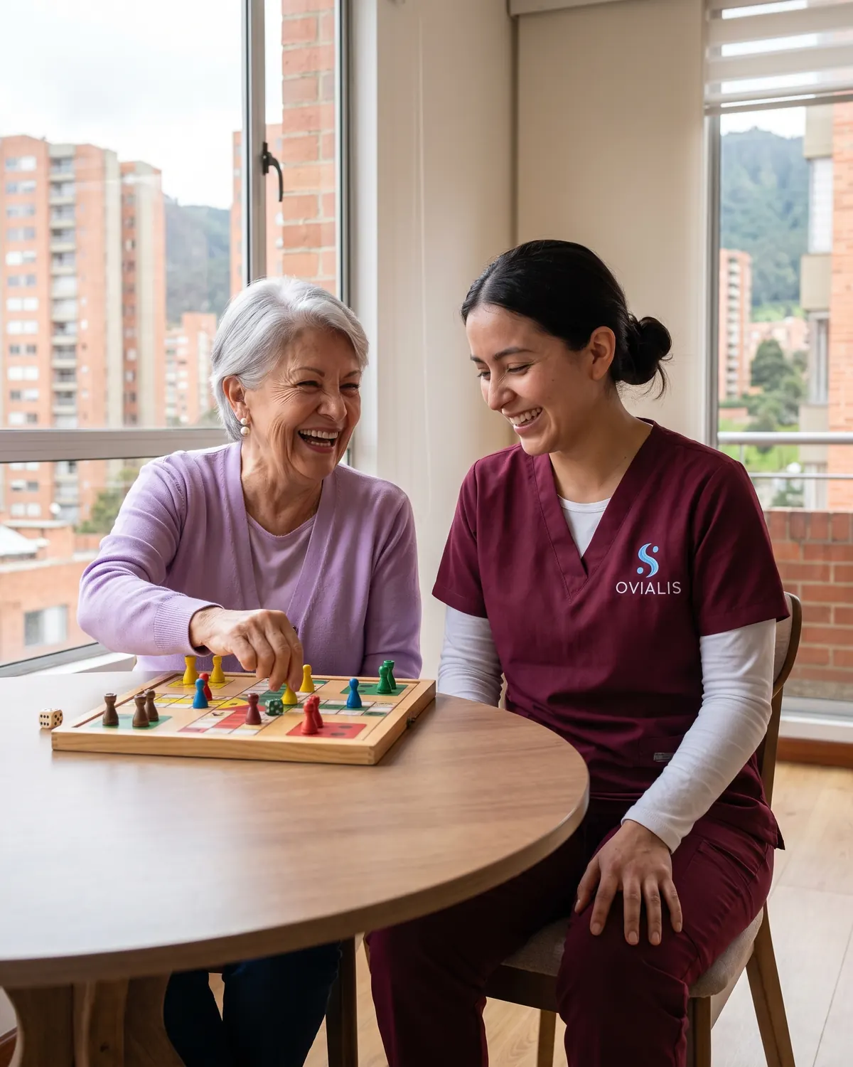 Cuidadora de adulto mayor y señora juegan parqués y se ríen junto a la ventana de un apartamento del norte de Bogotá